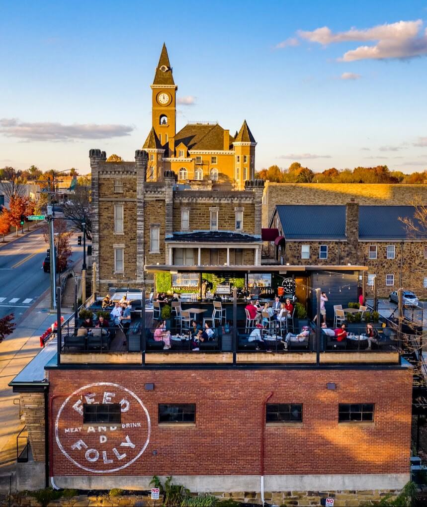 Rooftop restaurant with diners overlooks a historic brick building and clock tower in a charming SEC Town during autumn. Large "Feed & Folly" sign graces the side of the restaurant, adding to the small-town atmosphere.