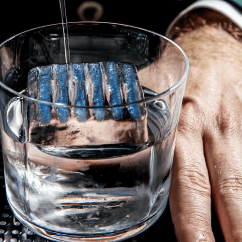 A close-up of a glass of clear liquid with a large, ridged blue ice cube, next to a person’s hand resting on a bar mat.