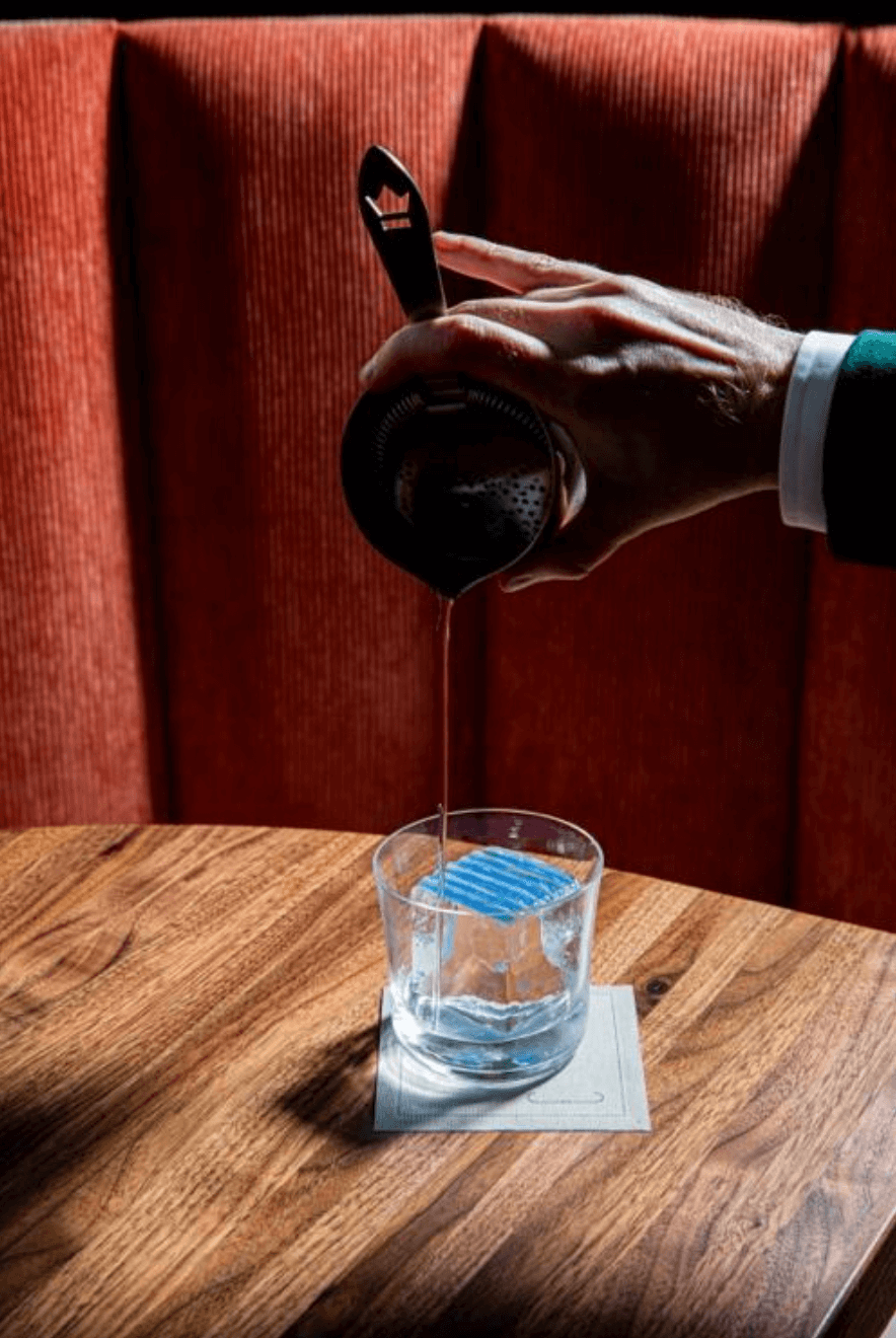 A hand pours a refreshing summer drink, the Catamaran Tan, from a cocktail shaker through a strainer into a glass on a napkin, set on a wooden table with a red upholstered background.