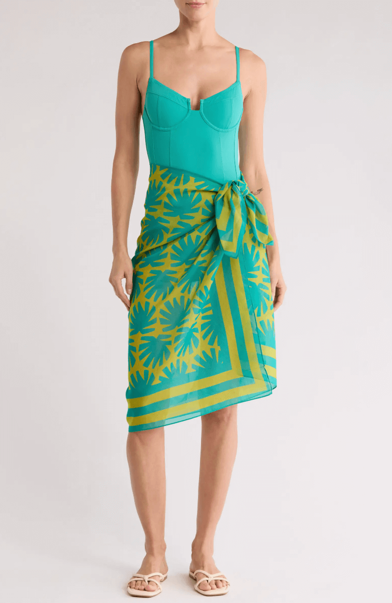 A woman wearing a teal fitted top, a green and yellow patterned wrap skirt tied at the waist, flat sandals, and subtle boating accessories stands against a plain background.