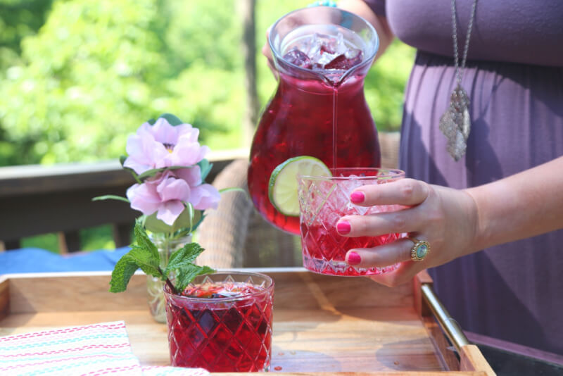 A person pours a red iced drink with lime slices from a pitcher into a glass on a wooden tray, skillfully arranged by Asheville artisans, next to a pink flower in a vase.