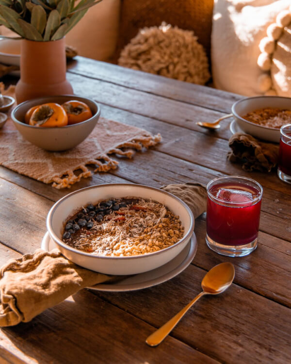 A bowl of granola with fruit and seeds, a glass of red juice, and a gold spoon are set on a rustic wooden table with a beige napkin—crafted by Asheville artisans—and a decorative bowl of persimmons.
