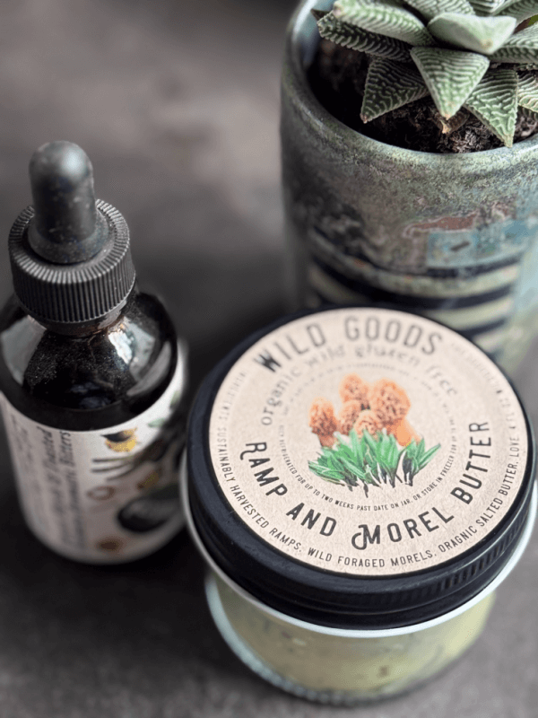 A jar labeled "Ramp and Morel Butter," crafted by Asheville artisans, sits next to a dropper bottle and a potted succulent on a dark surface.