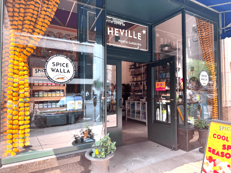 A storefront with a sign reading "Spice Walla" displays spice products crafted by Asheville artisans. The entrance is decorated with orange garlands, and an "OPEN" sign hangs on the door.