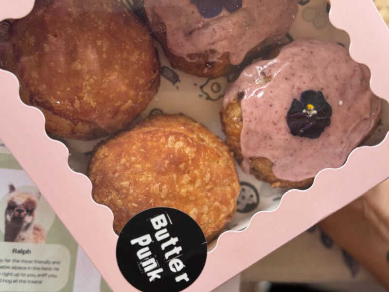 A box of four pastries—two plain, two with pink icing and flower decorations—labeled "Butter Punk," channels the creative flair often found at Asheville restaurants. A leaflet featuring an alpaca photo is partially visible.