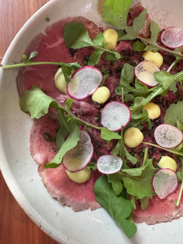 A plate of beef carpaccio topped with arugula, radish slices, yellow sauce dollops, and greens.