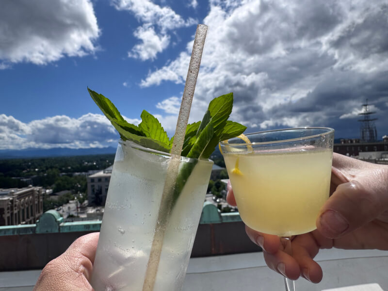 Two hands holding cocktails—one clear with mint and a straw, the other yellow with a twist—toast above Asheville restaurants on a city rooftop beneath a cloudy sky.
