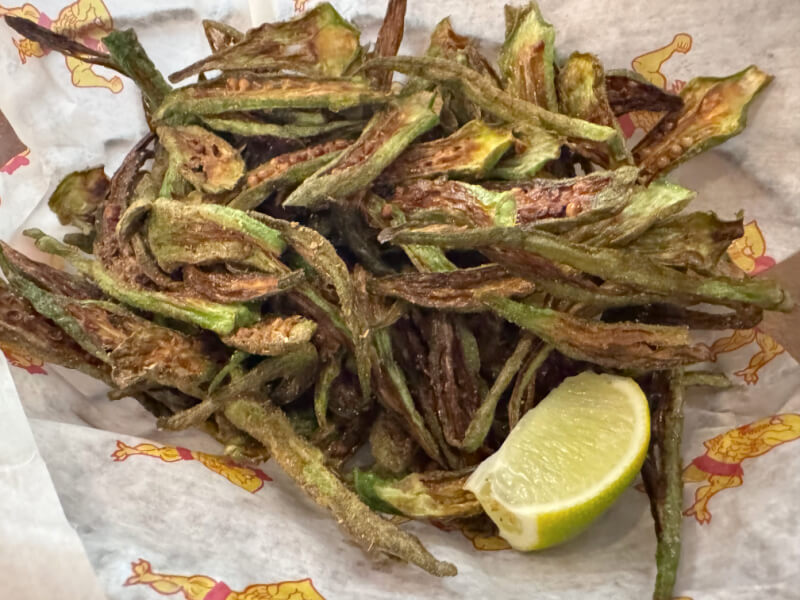 A basket of crispy fried okra strips, served with a wedge of lime on patterned paper.