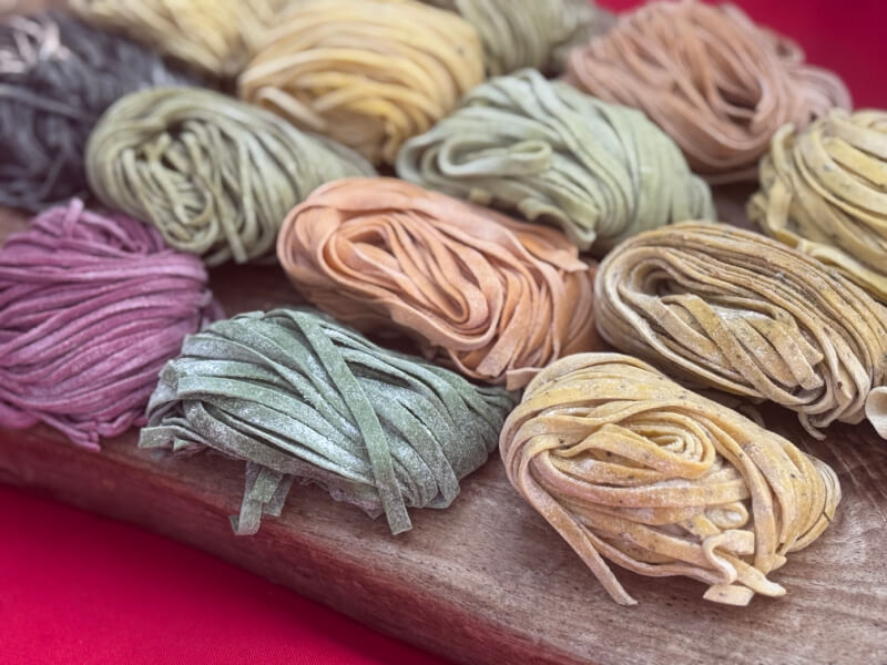 Assorted bundles of colorful uncooked pasta are arranged on a wooden board, with each bundle displaying a different natural hue.