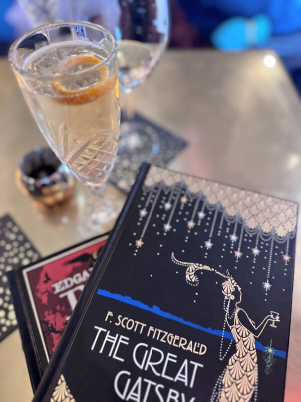 A glass of cocktail with a slice of orange sits beside a copy of "The Great Gatsby" by F. Scott Fitzgerald on a table, evoking the relaxed charm found in Asheville restaurants.