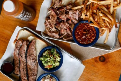 Two trays with barbecue brisket, pulled pork, French fries, baked beans, broccoli salad, and coleslaw sit on a wooden table. A bottle of barbecue sauce is also visible—perfect fare at restaurants open on July 4th.