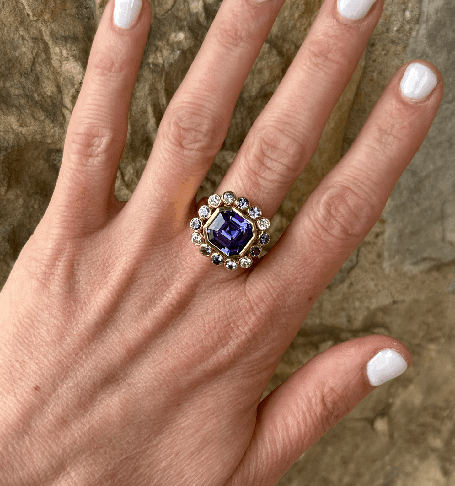 A hand with white nail polish wears a luxe gold ring featuring a large purple gemstone surrounded by clear stones, shown against a stone background—a stunning Birmingham find.