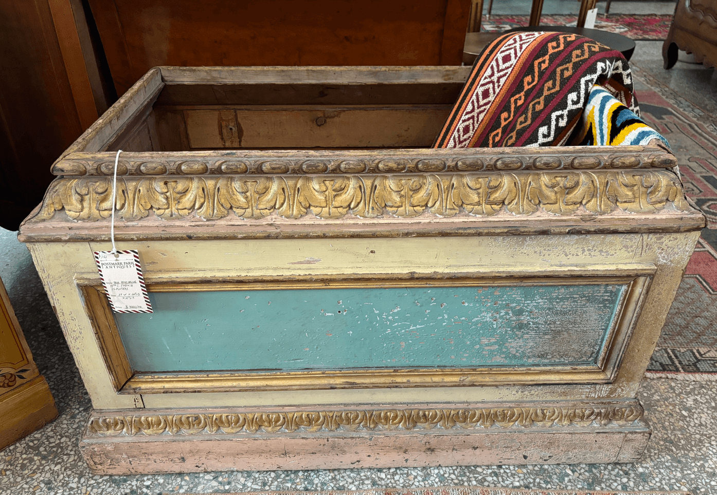 A vintage wooden chest with ornate carved trim and faded blue paneling, holding a patterned textile, is displayed indoors on a speckled floor—a luxe FINDS piece adding character to any Birmingham home.