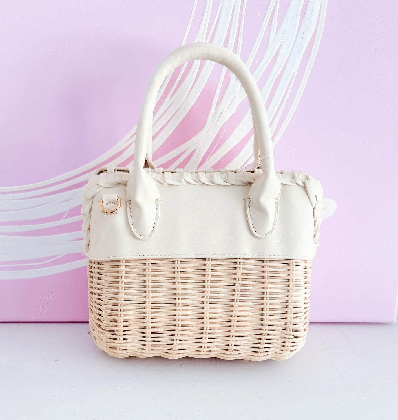 A small beige wicker handbag with white leather handles and trim, placed in front of a pink background with white curved lines.