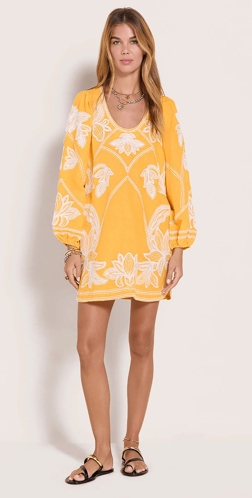 A woman stands against a plain background in a yellow, long-sleeved, floral-patterned mini dress with black sandals and layered necklaces—perfect for a sunny June day in Birmingham.