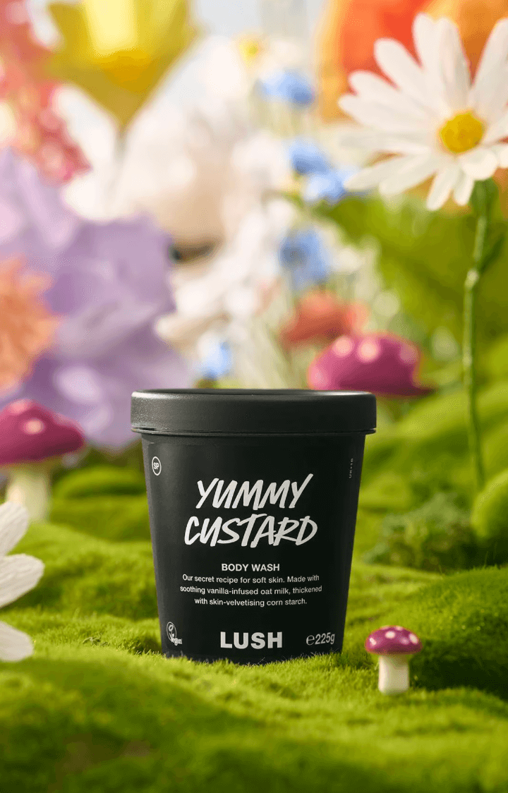 A black tub of Lush Yummy Custard body wash sits on green moss surrounded by artificial flowers and small purple mushrooms, evoking a whimsical Birmingham garden in June.