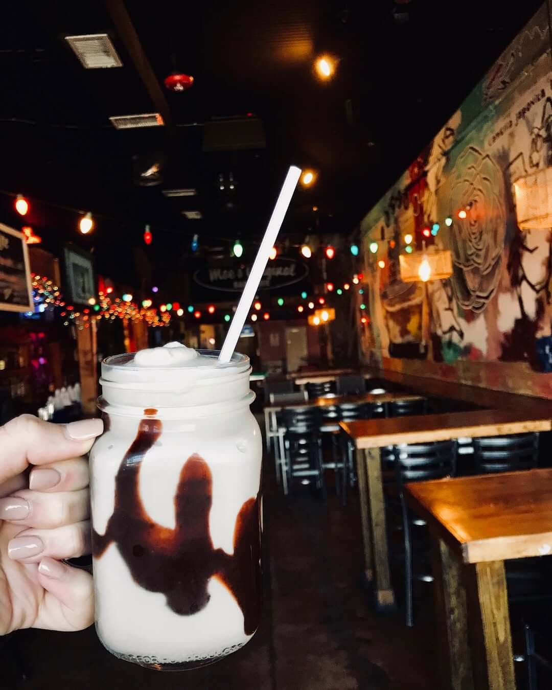 A hand holds a mason jar filled with a creamy Bushwacker frozen cocktail and chocolate syrup, topped with foam and a straw, inside Moe's BBQ.