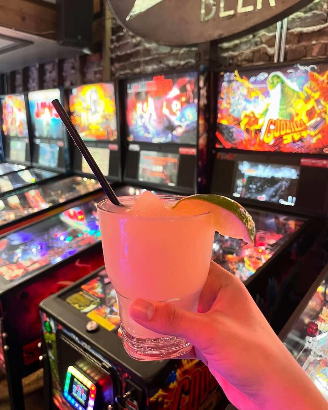 A hand holds a summer frozen cocktail with a lime wedge and straw in front of a row of colorful pinball machines at Paramount.