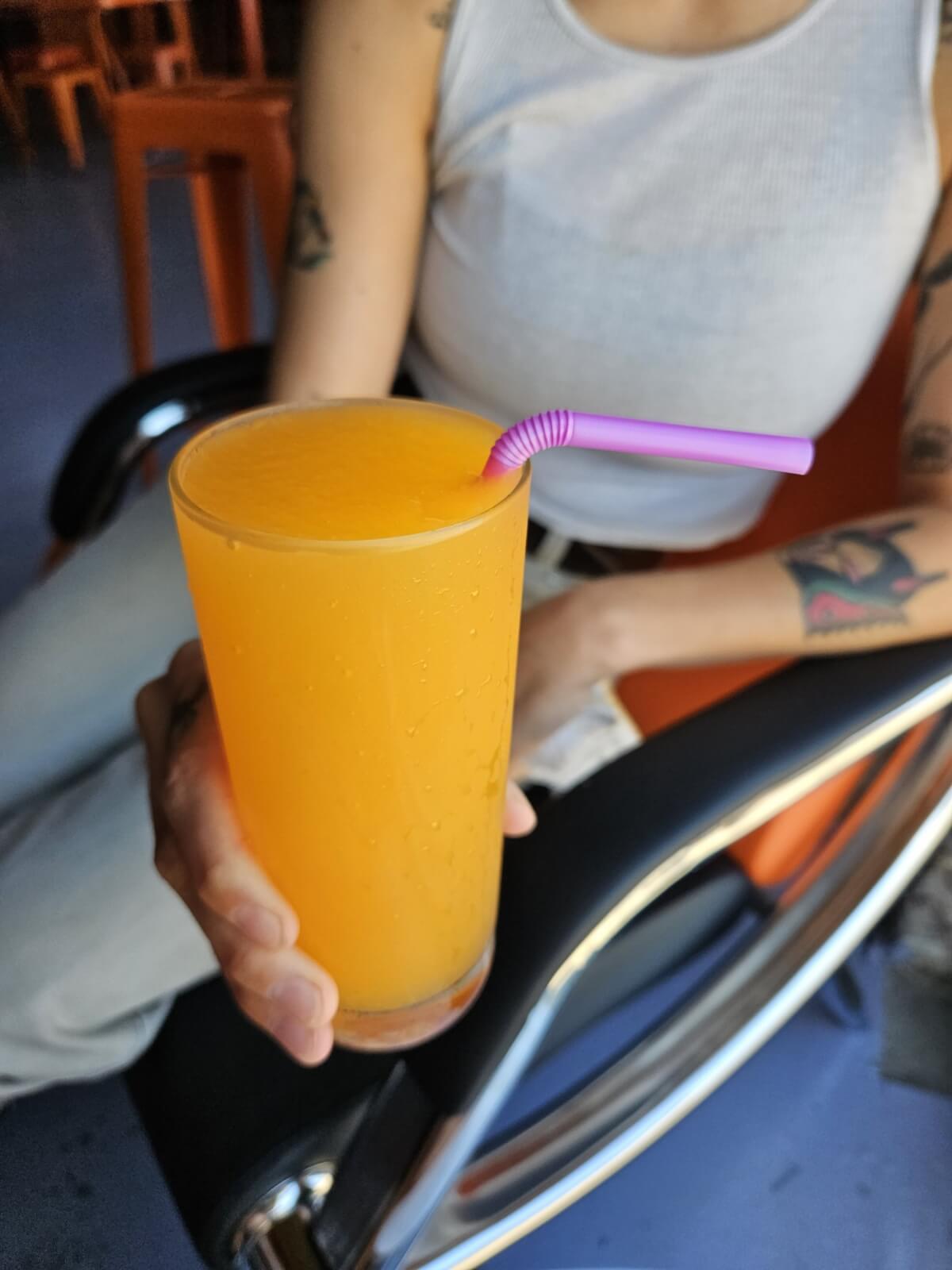 A person in a white tank top holds a frozen orange drink with a purple straw, perfect for enjoying summer vibes at Saturn + Satellite.