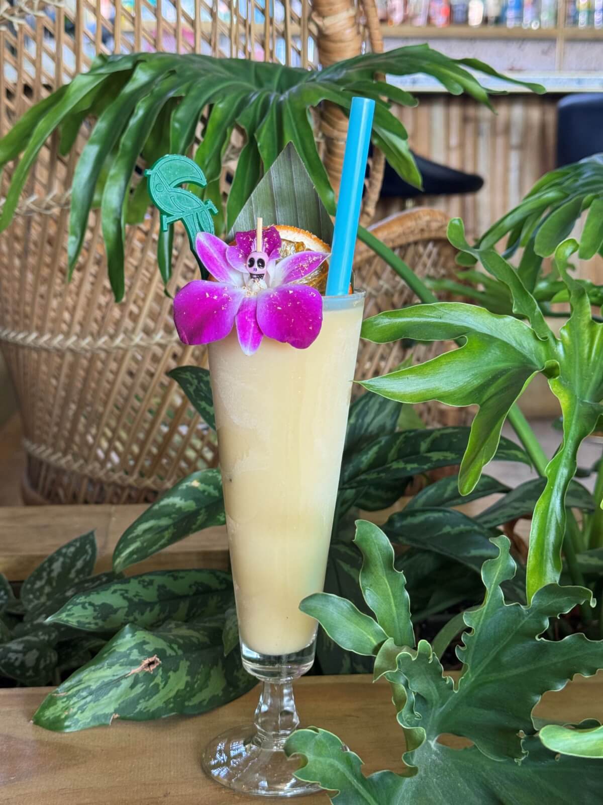A tall glass of Tucana's Frozen Painkiller: a yellow tropical frozen cocktail, garnished with a purple orchid, orange slice, green stir stick, and blue straw.