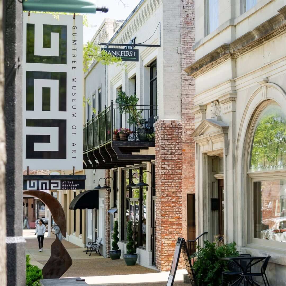 Street view of downtown Tupelo, MS, area with the Gumtree Museum of Art sign, perfect for weekend strolls or last-minute getaways from BHAM, featuring a person walking and inviting outdoor seating along the sidewalk.