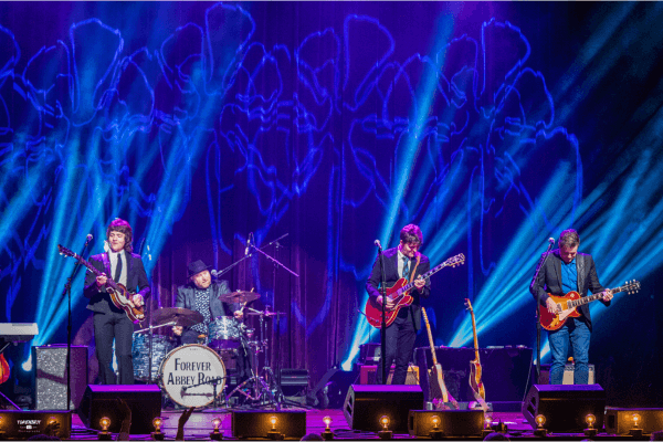 Four musicians perform on stage with guitars and drums under purple and blue lights at the Love Concert; a drum set displays the text "Forever Abbey Road.