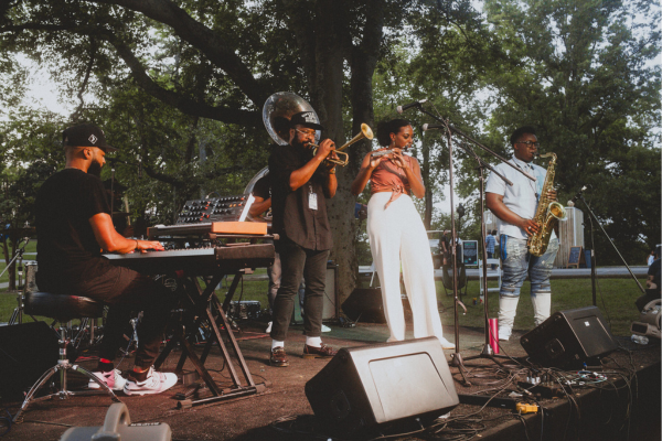 Four musicians perform on an outdoor stage at the Black Arts Bash with keyboard, sousaphone, trumpet, and saxophone, surrounded by trees and an engaged audience in the background.