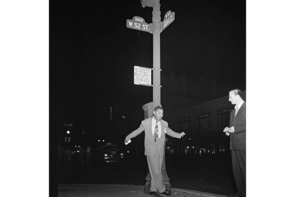 A man in a suit stands by a streetlamp at the corner of W. 52nd St. at night, facing another man in a dark suit—capturing the timeless atmosphere found in Classic Photographs from the Bank of America Collection.
