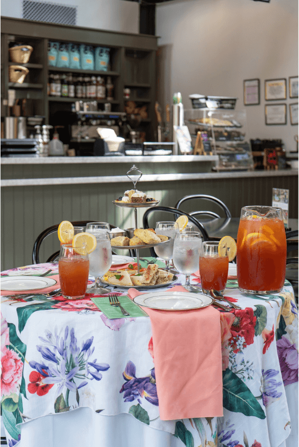 A floral-covered table set with iced tea, water, pastries, and sandwiches creates the perfect spot to book afternoon tea in Nashville, with a cozy café counter and shelves in the background.