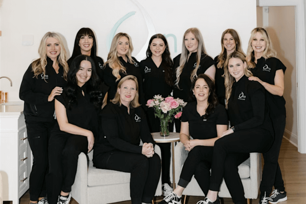 Group photo of eleven women in matching black uniforms, sitting and standing around a small table with a vase of pink flowers, celebrating Ona Skincare’s Grand Opening in the bright and welcoming Wellness East Nashville space.