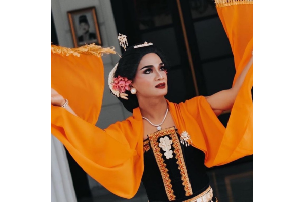 A person in traditional Indonesian attire with orange sleeves, floral hair accessories, and intricate jewelry performs a graceful Lengger Dance with raised arms during a studio presentation.