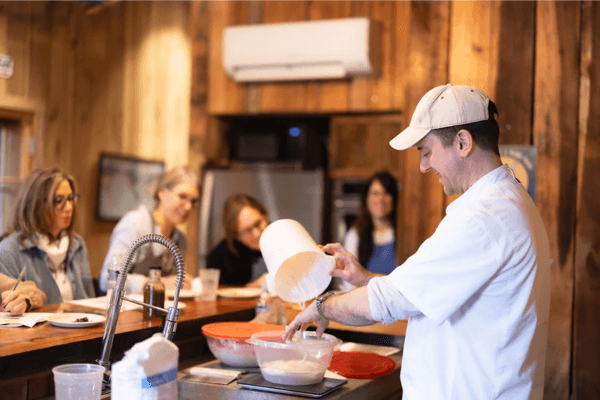 A man in a white shirt and cap demonstrates mixing ingredients in a bowl to a group of seated women during a Sourdough Bread Making Class at West Glow Farms, set in a rustic kitchen on a bright morning & afternoon.