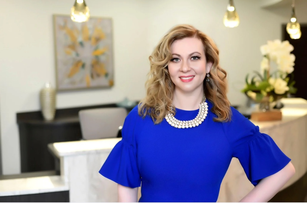 A woman with wavy blonde hair, wearing a royal blue dress and pearl necklace, stands smiling in a modern office at Cahaba Dermatology, with artwork and flowers in the background.