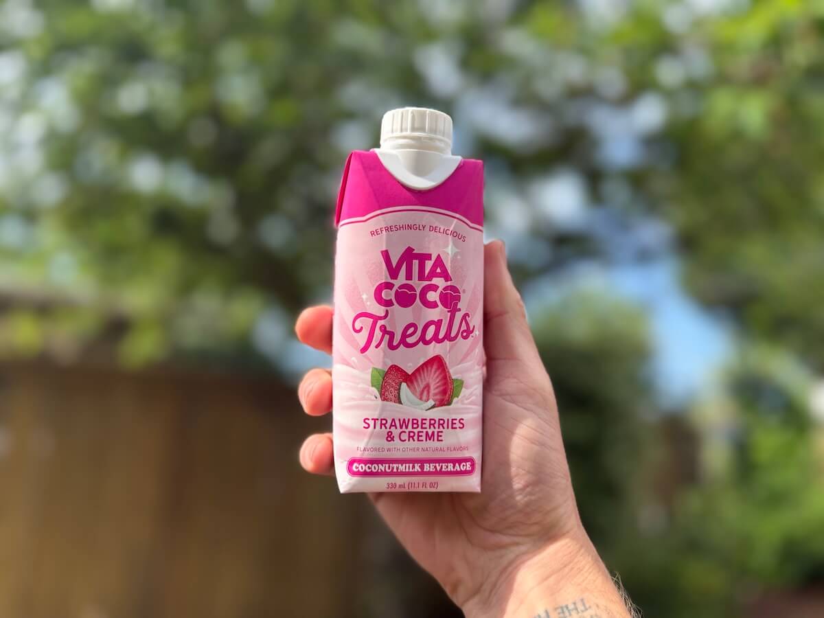 A hand holds a carton of Vita Coco Treats Strawberries & Creme coconutmilk beverage—one of the summer essentials—against a blurred outdoor background.