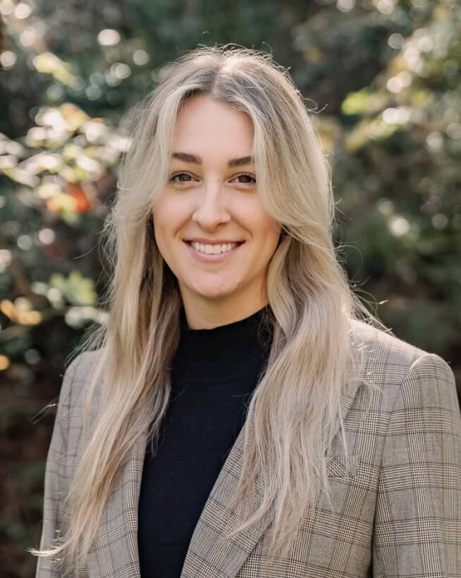 A woman with long blonde hair, wearing a plaid blazer and black top, stands outdoors in front of blurred greenery, smiling at the camera.