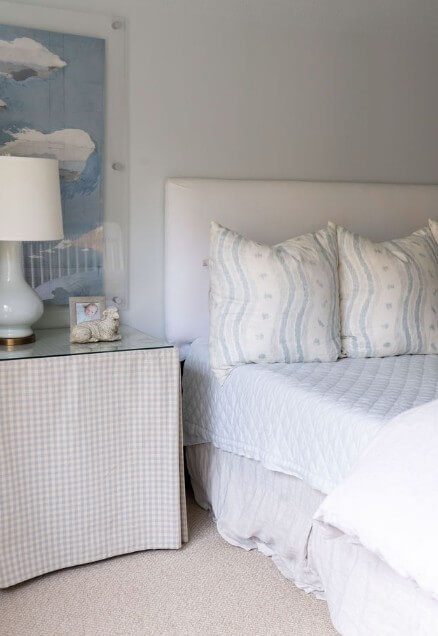 A neatly made bed with white and light blue pillows sits beside a table covered with a checkered cloth, styled by Logos Interiors, featuring a lamp, photo frame, and small decorative item.
