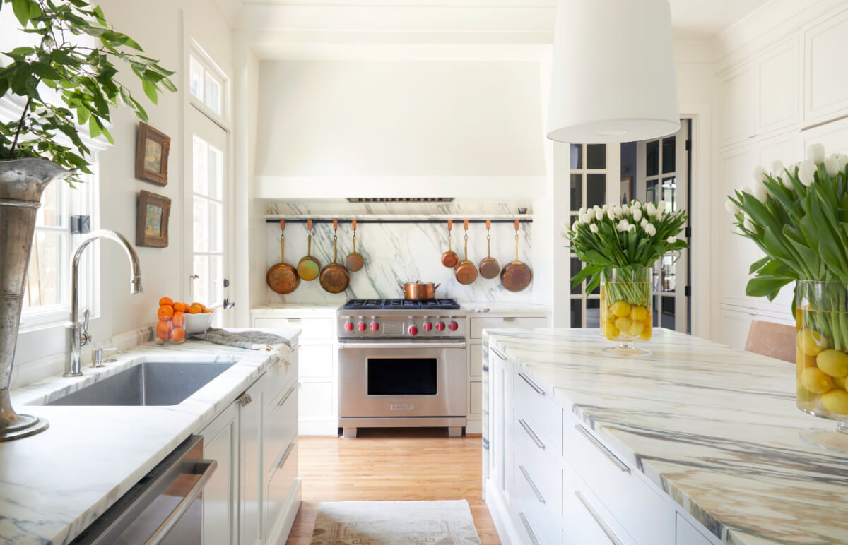 Bright, modern kitchen by Logos Interiors with marble countertops, stainless steel stove, copper pots hanging above, and vases of lemons and tulips as decoration.