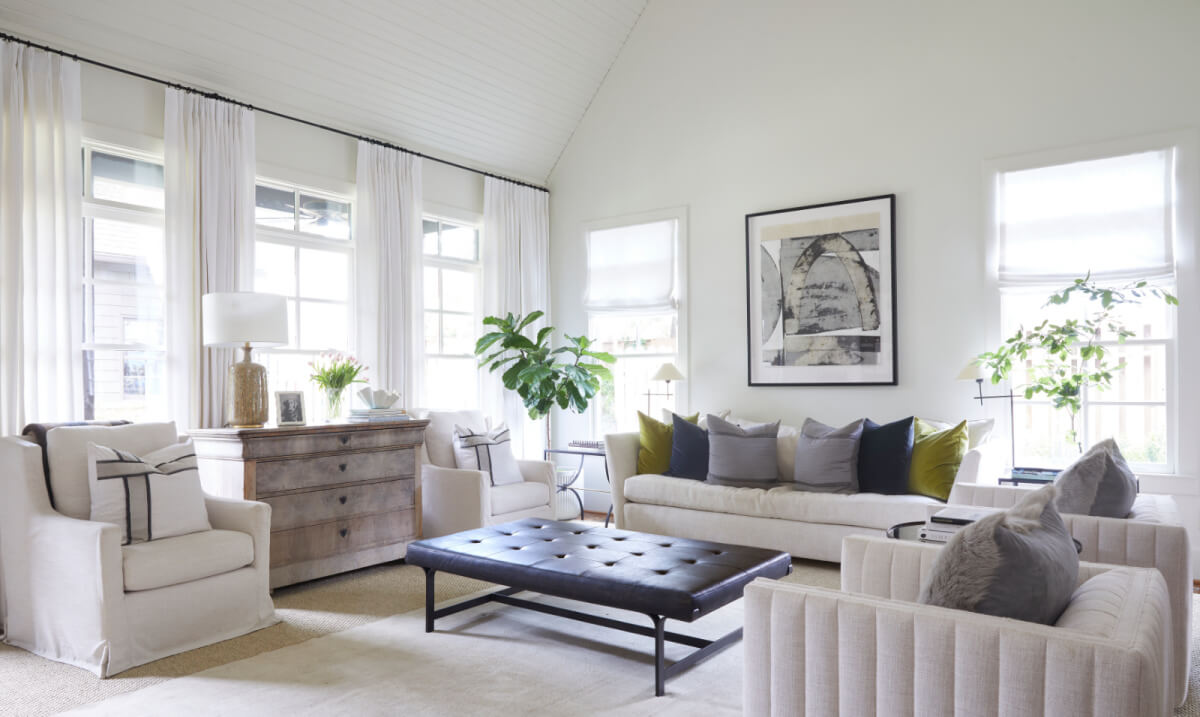 Bright living room by Logos Interiors with white walls, neutral furniture, large windows, sheer curtains, plants, a wooden chest, modern artwork, and a black tufted bench table in the center.