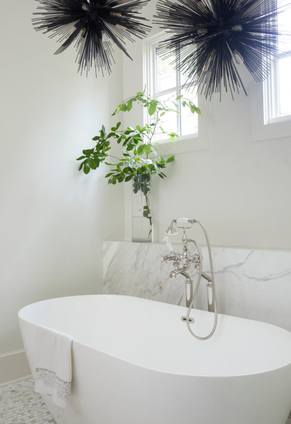 A modern white bathtub with a towel draped over the edge sits below two spiky black light fixtures and a window with a leafy plant on the ledge, showcasing the sleek style of Logos Interiors.
