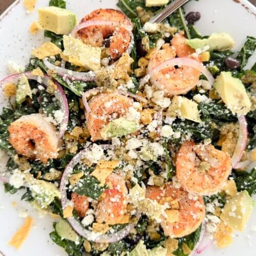 A plate of Shrimp Kale Salad with avocado, black beans, red onions, crumbled cheese, tortilla chips, and a zesty Lime Pepita Dressing sits on a wooden table.