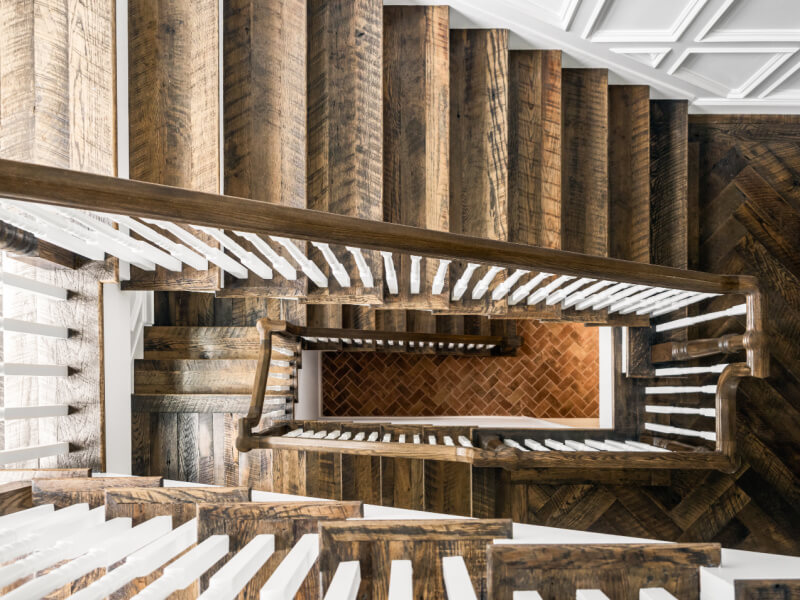 View from above of a wooden staircase with white spindles, descending multiple levels to a landing with a herringbone-patterned brick floor..