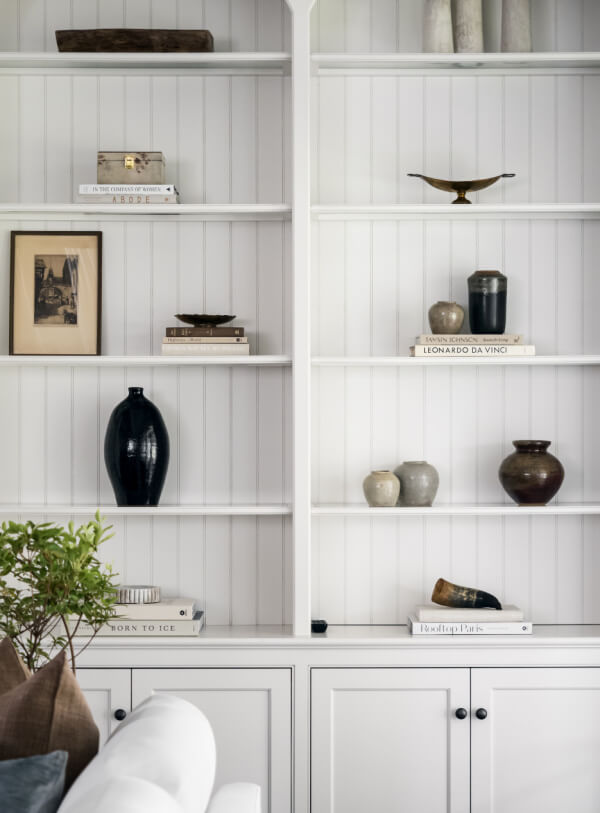 White built-in shelves display books, vases, pottery, a framed photo, and a decorative bowl—a stylish home feature. A green potted plant sits in front of the shelves.