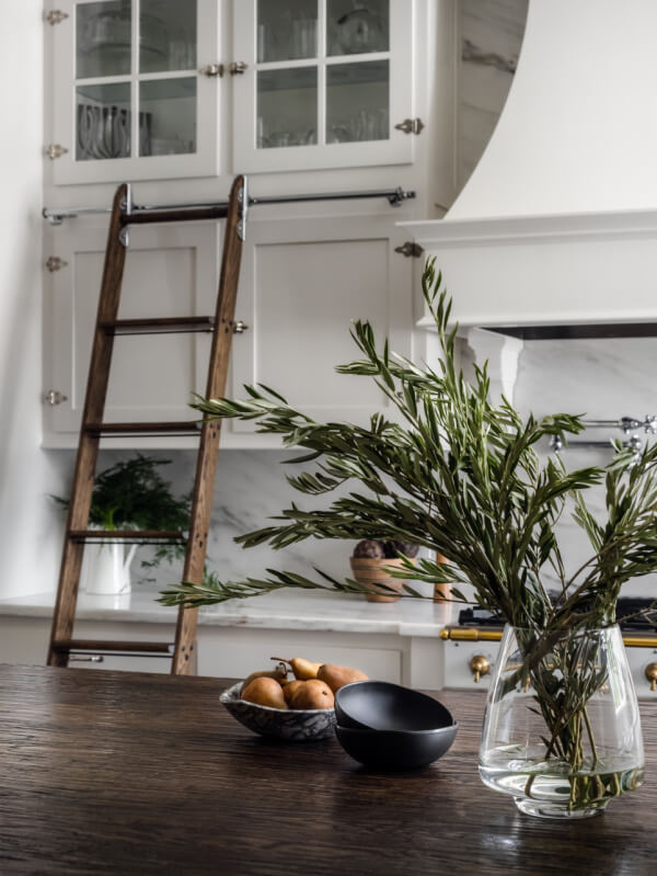 A kitchen scene in a Georgia home features a wooden ladder against white cabinets, a glass vase with greenery, and bowls of fruit on a dark wooden table.