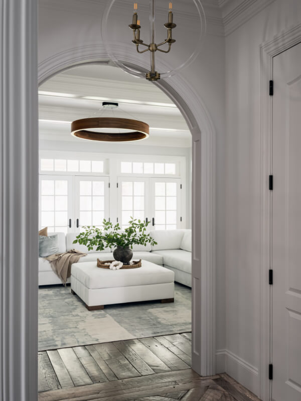A view through an arched doorway into a bright living room with a white sectional sofa, large ottoman, potted plant, and modern light fixtures.