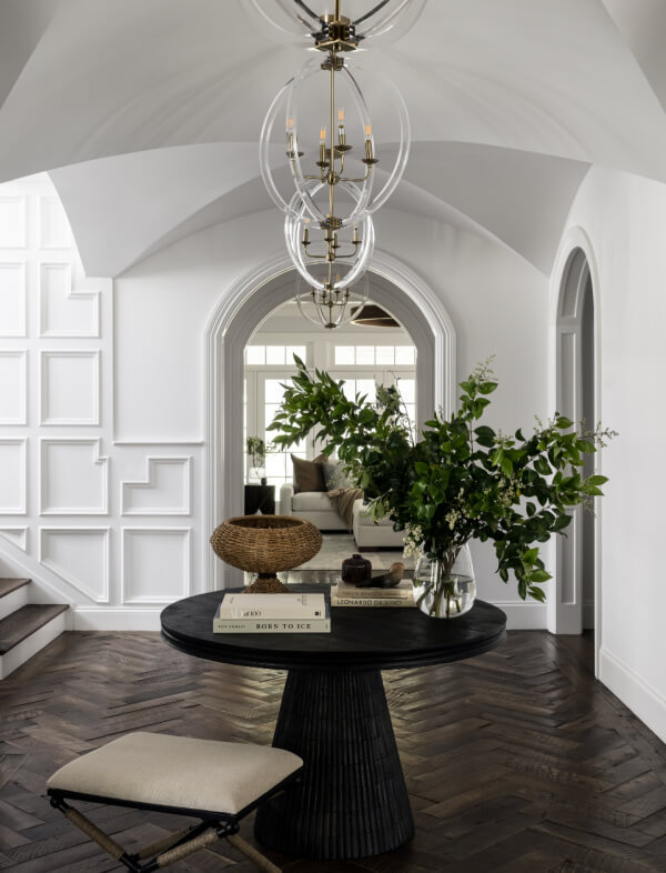 A round black table with books and greenery sits in a white entryway with herringbone wood floors, arched doorways, and a modern chandelier.