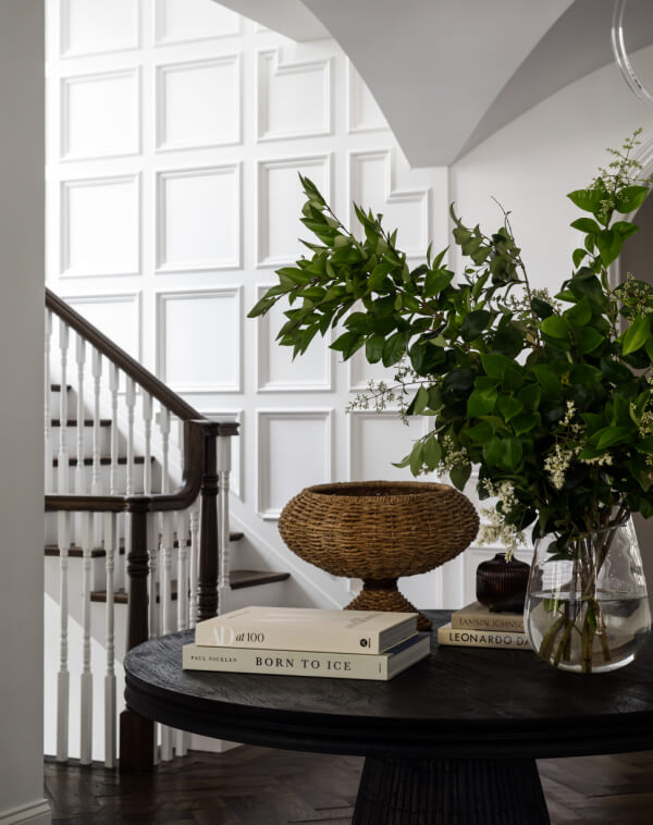 A round black table with books, a wicker basket, and a glass vase with green branches sits in a hallway near a staircase with white paneled walls and dark wood accents.