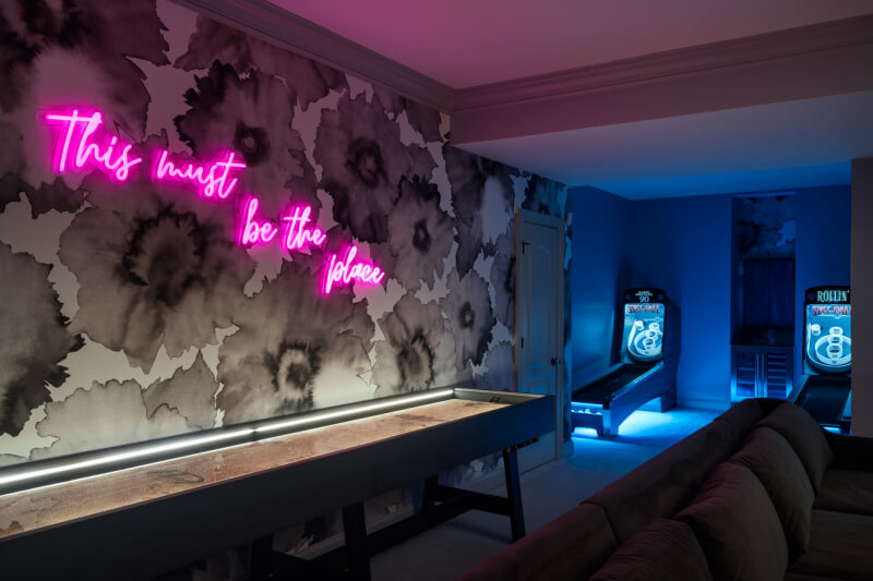 A room with floral wallpaper, a neon sign reading "This must be the place," shuffleboard table, couch, and two skee-ball arcade machines in the background.