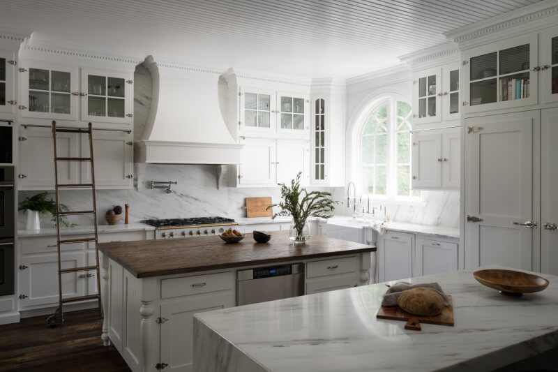 A spacious white Georgia & Hunt kitchen features marble countertops, a wooden island, glass-front cabinets, a large range hood, and an arched window letting in natural light.