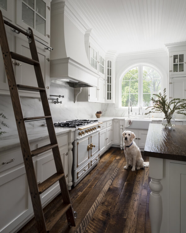A white Georgia & Hunt kitchen with a farmhouse sink, wood flooring, a rolling ladder, and a golden retriever sitting near the island. Large arched window lets in natural light.