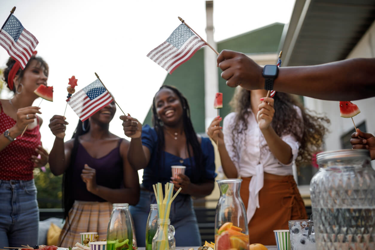 A group of people holding small American flags and watermelon slices stand around a table with snacks and drinks at an outdoor gathering, celebrating one of the lively events in Louisville.